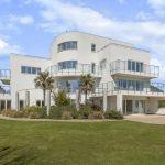 Property for sale by Auction A modern, multi-story white house with extensive glass panel balconies and large windows, set in a well-maintained garden on Dilks Street. The house features several rounded and angular architectural elements and is surrounded by lush green grass and a few palm trees. The sky is partly cloudy. Presented by Palace Auctions
