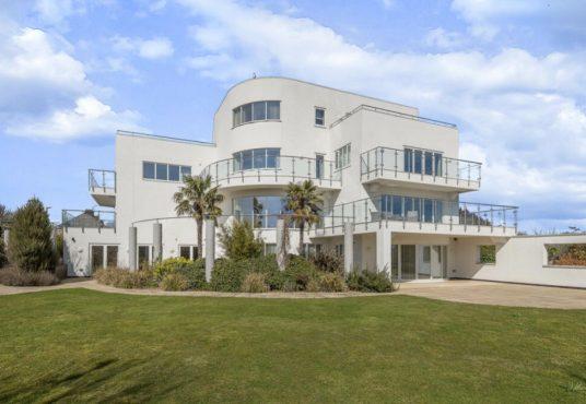 Property for sale by Auction A modern, multi-story white house with extensive glass panel balconies and large windows, set in a well-maintained garden on Dilks Street. The house features several rounded and angular architectural elements and is surrounded by lush green grass and a few palm trees. The sky is partly cloudy. Presented by Palace Auctions