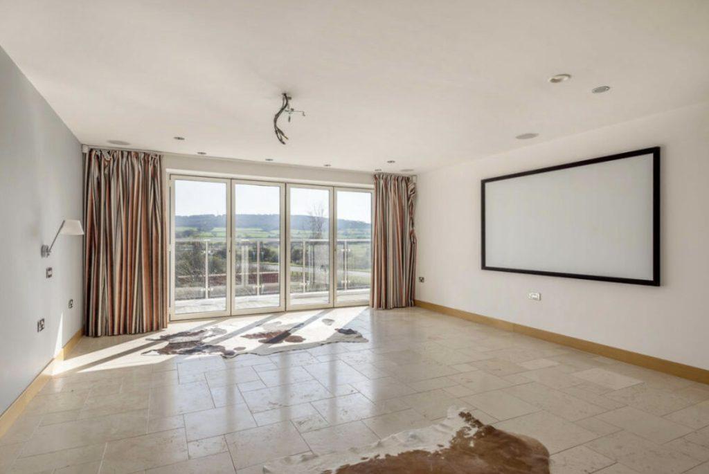 Property for sale by Auction A spacious, empty room with large floor-to-ceiling windows framed by curtains on the left and right, offering a view of the scenic Tindale Towers. The room has light-colored tile flooring, a blank wall-mounted rectangular screen, and two animal hide rugs on the floor. Presented by Palace Auctions