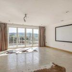 Property for sale by Auction A spacious, empty room with large floor-to-ceiling windows framed by curtains on the left and right, offering a view of the scenic Tindale Towers. The room has light-colored tile flooring, a blank wall-mounted rectangular screen, and two animal hide rugs on the floor. Presented by Palace Auctions