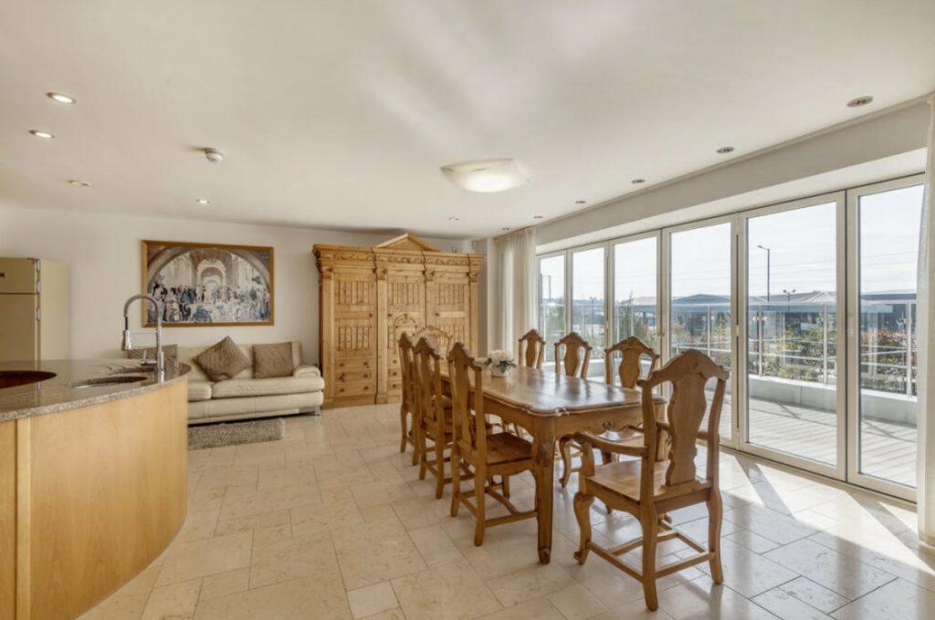 Property for sale by Auction A spacious, modern dining room at Tindale Towers on Dilks Street features a large wooden table with eight matching chairs. Floor-to-ceiling windows offer a scenic view. To the left is a curved kitchen countertop with a sink, while a beige couch and ornate wooden cabinet adorn the back wall of this Bishop DL14 9AP gem. Presented by Palace Auctions