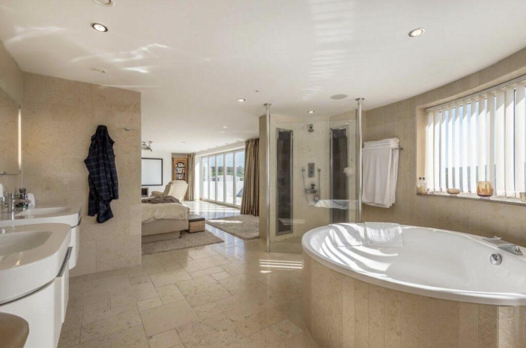 Property for sale by Auction Spacious bathroom with a large oval bathtub, a glass shower enclosure, double sinks, and neutral beige tiles. A bathrobe hangs by the sinks. The room extends into a bedroom, allowing natural light to fill both spaces through windows with blinds—perfect for a home on Dilks Street. Presented by Palace Auctions