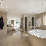 Property for sale by Auction Spacious bathroom with a large oval bathtub, a glass shower enclosure, double sinks, and neutral beige tiles. A bathrobe hangs by the sinks. The room extends into a bedroom, allowing natural light to fill both spaces through windows with blinds—perfect for a home on Dilks Street. Presented by Palace Auctions