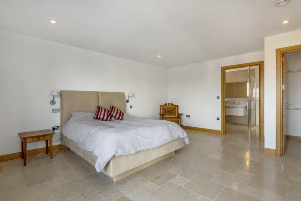 Property for sale by Auction A spacious bedroom, located in Tindale Towers on Dilks Street, boasts a large beige bed adorned with striped pillows. The room features light-colored tiled flooring, a wooden nightstand, and a wooden dresser. A door leads to an attached bathroom with a sink and large mirror visible inside. Walls are painted white. Presented by Palace Auctions