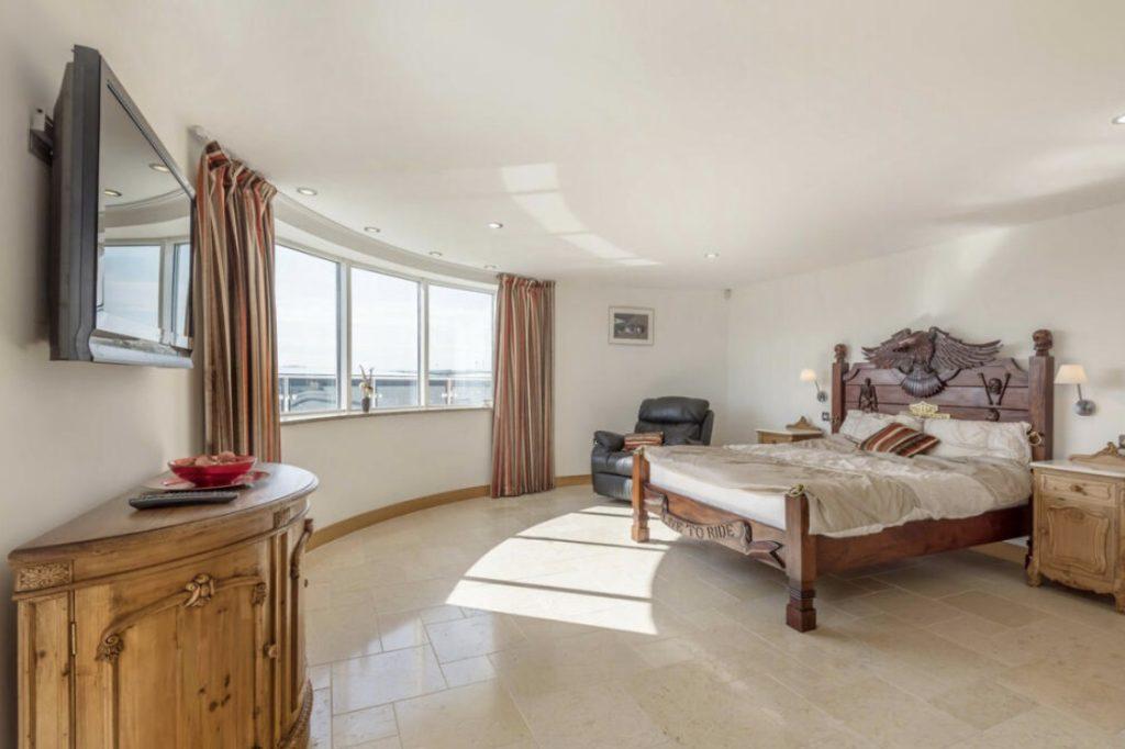 Property for sale by Auction A spacious bedroom at Tindale Towers, Bishop DL14 9AP, with a large curved window covered by red curtains. It features a wooden bed with an eagle-themed headboard, a leather recliner chair, a wall-mounted TV, a wooden dresser with a decorative bowl, and a nightstand with a lamp and picture above it. Presented by Palace Auctions
