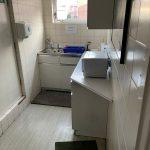 Property for sale by Auction A small white-tiled kitchenette with a sink, faucet, and blue dish rack under the window. Above the counter is a cabinet. On the counter beside the sink is a microwave. There are soap dispensers and warning signs on the wall—an ideal feature in this Church Walk pharmacy investment property available for sale by auction. Presented by Palace Auctions