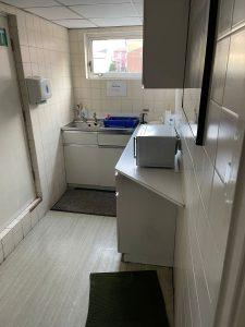 Property for sale by Auction A small white-tiled kitchenette with a sink, faucet, and blue dish rack under the window. Above the counter is a cabinet. On the counter beside the sink is a microwave. There are soap dispensers and warning signs on the wall—an ideal feature in this Church Walk pharmacy investment property available for sale by auction. Presented by Palace Auctions