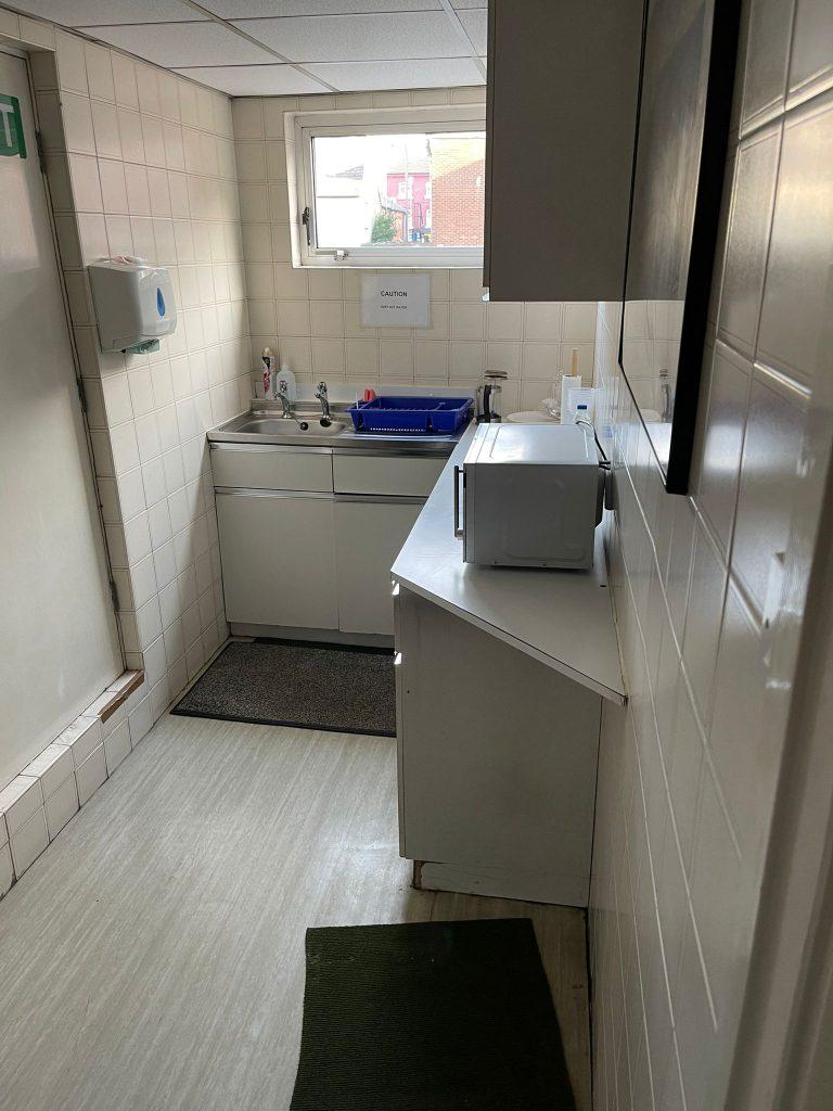 Property for sale by Auction A small white-tiled kitchenette with a sink, faucet, and blue dish rack under the window. Above the counter is a cabinet. On the counter beside the sink is a microwave. There are soap dispensers and warning signs on the wall—an ideal feature in this Church Walk pharmacy investment property available for sale by auction. Presented by Palace Auctions