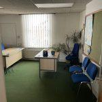 Property for sale by Auction A small office with a green carpet, white walls, and a large window with vertical blinds. The room has a desk with office supplies, two blue chairs, and a whiteboard. Several potted plants are placed around the room. There's also a notice board on the wall announcing an investment property available for sale by auction. Presented by Palace Auctions