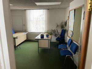 Property for sale by Auction A small office with a green carpet, white walls, and a large window with vertical blinds. The room has a desk with office supplies, two blue chairs, and a whiteboard. Several potted plants are placed around the room. There's also a notice board on the wall announcing an investment property available for sale by auction. Presented by Palace Auctions