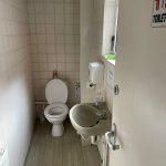 Property for sale by Auction A small restroom with a white toilet and open lid, a beige sink, and a liquid soap dispenser mounted on the wall. A small window sits above the sink, providing natural light. The door has a "Toilet" sign and is partially open, revealing tiled walls and a radiator in this Church Walk pharmacy investment property available for sale by auction. Presented by Palace Auctions