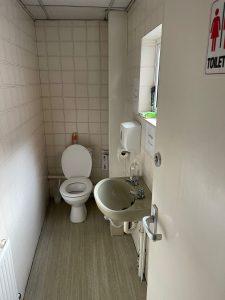 Property for sale by Auction A small restroom with a white toilet and open lid, a beige sink, and a liquid soap dispenser mounted on the wall. A small window sits above the sink, providing natural light. The door has a "Toilet" sign and is partially open, revealing tiled walls and a radiator in this Church Walk pharmacy investment property available for sale by auction. Presented by Palace Auctions