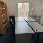 Property for sale by Auction A small office with two white desks forming an L-shape, two black office chairs, a wooden bookshelf on the left, a white wall cabinet on the right, and a window with vertical blinds letting in natural light. The room features blue carpeting and white walls—located near Church Walk Pharmacy. Presented by Palace Auctions