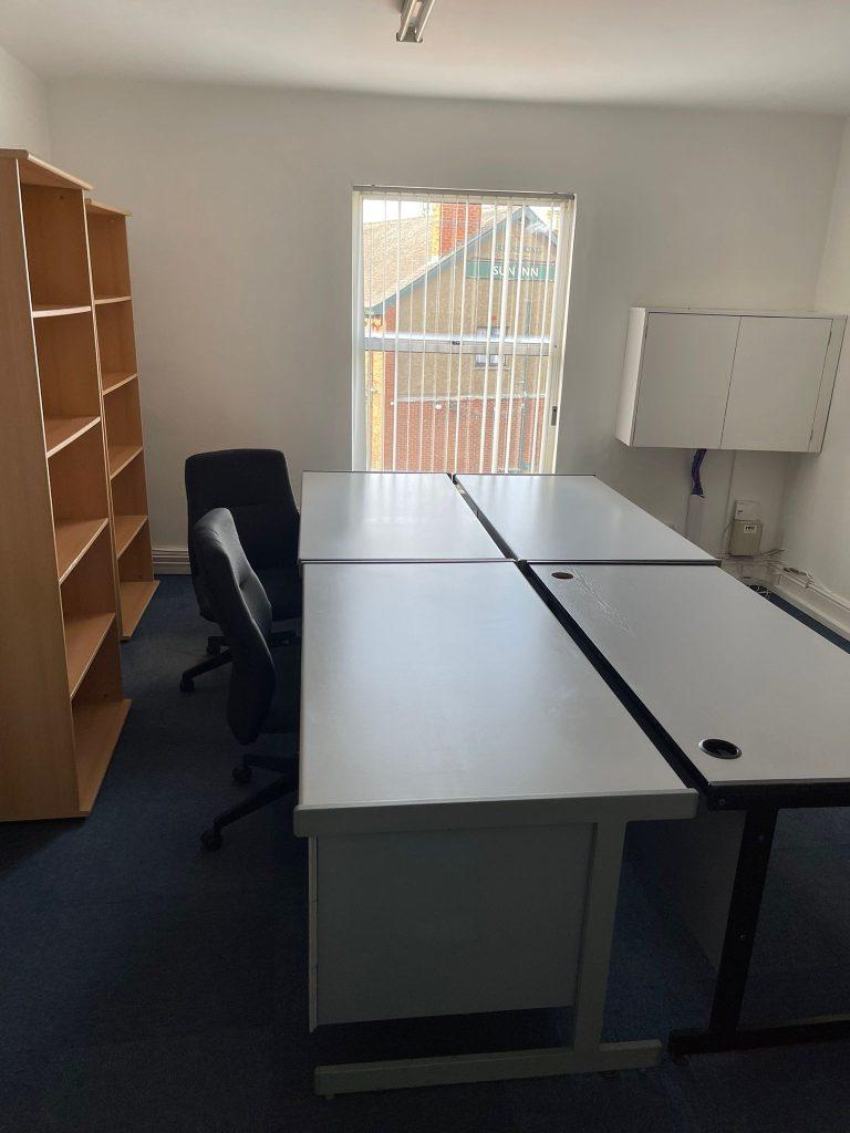 Property for sale by Auction A small office with two white desks forming an L-shape, two black office chairs, a wooden bookshelf on the left, a white wall cabinet on the right, and a window with vertical blinds letting in natural light. The room features blue carpeting and white walls—located near Church Walk Pharmacy. Presented by Palace Auctions