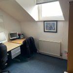 Property for sale by Auction A small attic office with a skylight. The room has a desk with a computer monitor, keyboard, and stacks of papers on it. A black jacket is draped over a chair next to the desk. A framed picture hangs above the radiator, and there's blue carpet on the floor—much like the charm seen in Church Walk properties available for sale by auction. Presented by Palace Auctions