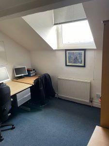 Property for sale by Auction A small attic office with a skylight. The room has a desk with a computer monitor, keyboard, and stacks of papers on it. A black jacket is draped over a chair next to the desk. A framed picture hangs above the radiator, and there's blue carpet on the floor—much like the charm seen in Church Walk properties available for sale by auction. Presented by Palace Auctions