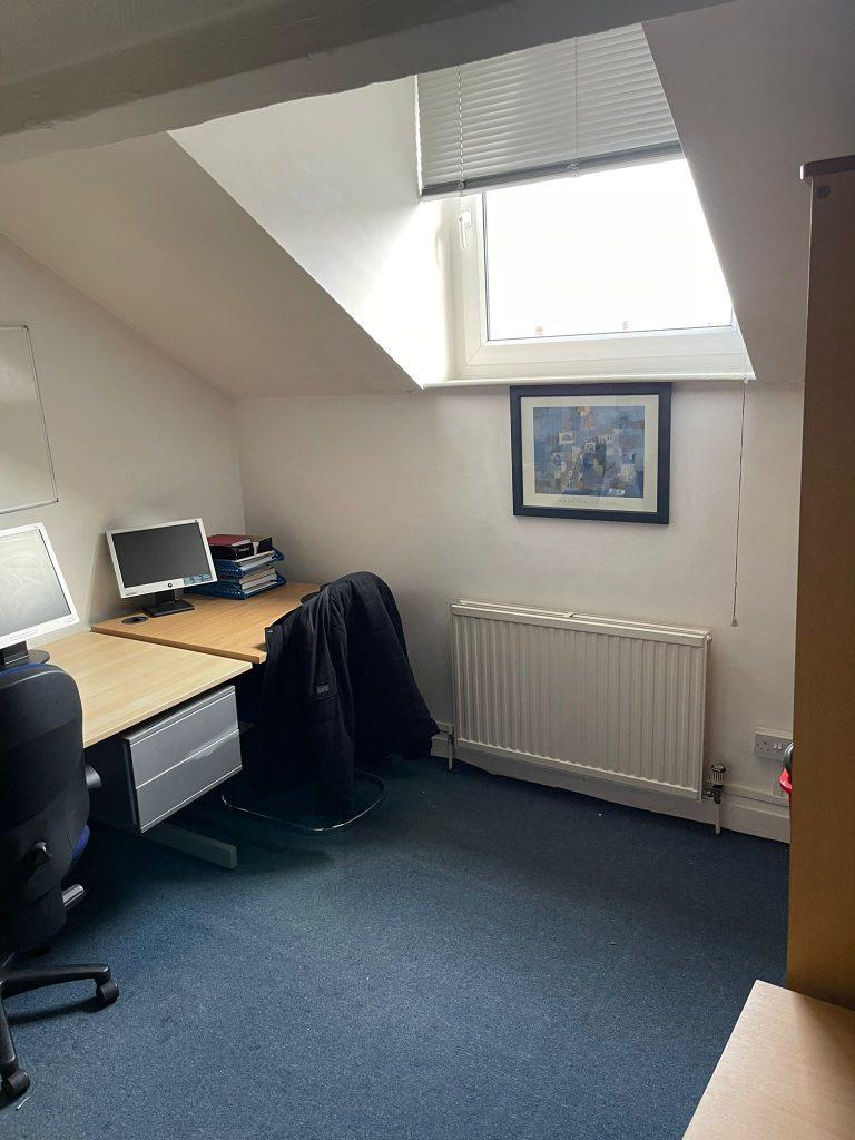 Property for sale by Auction A small attic office with a skylight. The room has a desk with a computer monitor, keyboard, and stacks of papers on it. A black jacket is draped over a chair next to the desk. A framed picture hangs above the radiator, and there's blue carpet on the floor—much like the charm seen in Church Walk properties available for sale by auction. Presented by Palace Auctions