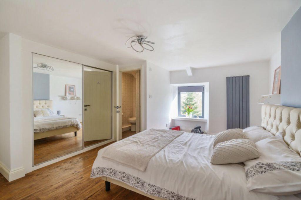 Property for sale by Auction A cozy bedroom at Hagg Hill Farm with a large bed featuring patterned pillows and a beige headboard. The room has a wooden floor and a mirrored wardrobe on the left. There's a small window with a radiator underneath it and a door to an en-suite bathroom. The decor is minimalist. Presented by Palace Auctions