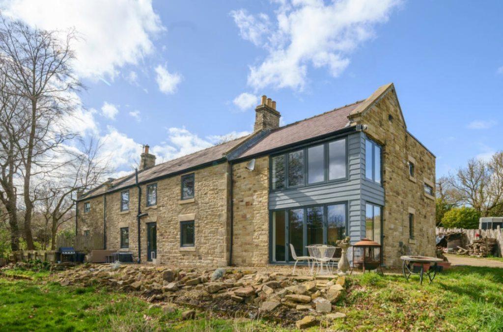 Property for sale by Auction A large stone house on Hagg Hill Farm boasts multiple chimney stacks and a mix of traditional and modern architectural elements. The house features large windows and a spacious patio area with outdoor seating, set against a backdrop of trees and a partly cloudy sky. Presented by Palace Auctions