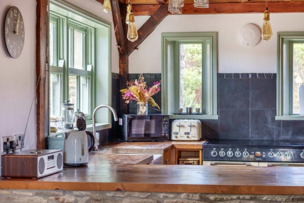 Property for sale by Auction A cozy kitchen at Hagg Hill Farm featuring a rustic wooden countertop, green-framed windows, and various appliances including a microwave, kettle, toaster, and radio. A vase of colorful flowers sits on the counter, while pots hang overhead. The room is well-lit by natural light. Presented by Palace Auctions