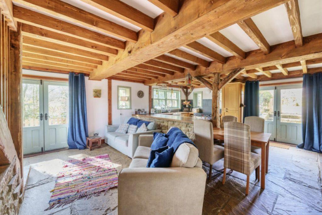Property for sale by Auction A cozy living space at Hagg Hill Farm with exposed wooden beams and rustic stone flooring. It features a beige sofa adorned with blue cushions, a colorful woven rug, wooden dining table with upholstered chairs, and multiple glass doors letting in natural light. Curtains are drawn open. Presented by Palace Auctions