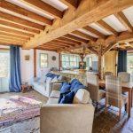 Property for sale by Auction A cozy living space at Hagg Hill Farm with exposed wooden beams and rustic stone flooring. It features a beige sofa adorned with blue cushions, a colorful woven rug, wooden dining table with upholstered chairs, and multiple glass doors letting in natural light. Curtains are drawn open. Presented by Palace Auctions