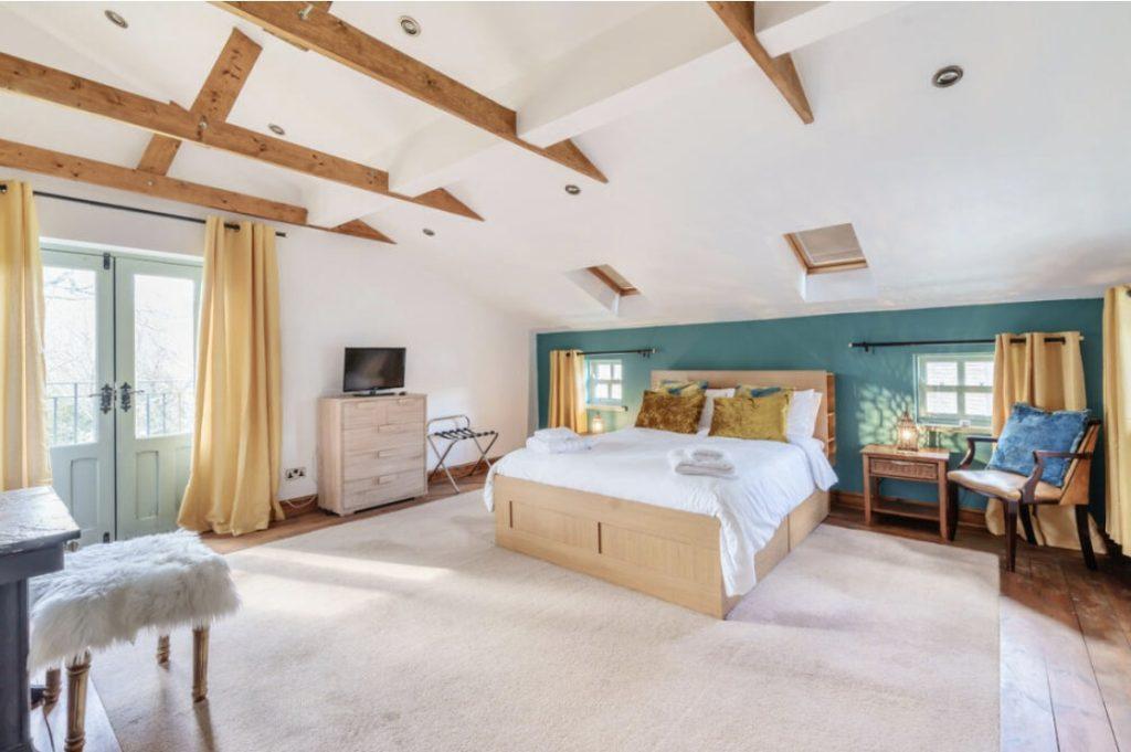 Property for sale by Auction A spacious bedroom at Hagg Hill Farm features wooden beams on the ceiling, a large bed with white linens and yellow and green accent pillows, a dresser with a TV, and an armchair. Light pours in through skylights and windows with yellow curtains. A fluffy stool sits beside a small table. Presented by Palace Auctions