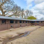 Property for sale by Auction A row of wooden stables with partially open upper doors lines up against a concrete pathway at Hagg Hill Farm. Water puddles dot the ground, and leafless trees stand in the background under a cloudy sky. The stables, with their black roofs, create a charming scene in this rural, outdoor setting. Presented by Palace Auctions