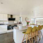 Property for sale by Auction A modern kitchen with white cabinets and a large island featuring a quartz countertop and four yellow bar stools from Hagg Hill Farm. The kitchen includes stainless steel appliances, a black range hood, and a coffee maker. A large clock hangs on a wall near floor-to-ceiling windows with a scenic view. Presented by Palace Auctions