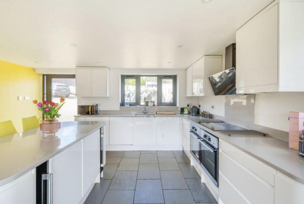 Property for sale by Auction A modern kitchen featuring white cabinets, gray countertops, and a sleek design. There's a built-in oven and stovetop on the right, a sink centered under a window at the far end, and a vase of tulips on the left counter. A bright yellow wall adds a pop of color. Presented by Palace Auctions