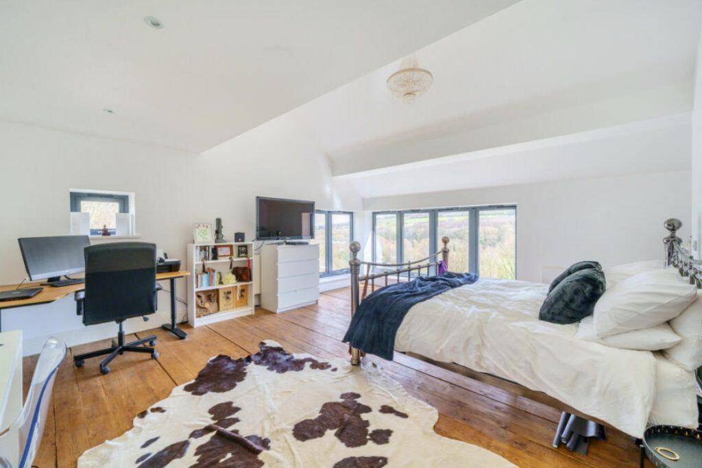 Property for sale by Auction A bright bedroom features a large window with scenic views, a bed with metal frame and white linens, a cowhide rug on wooden floors, and a workspace with a desk, computer, and office chair. Opposite the bed is a white dresser with a TV on top and storage shelves. The decor hints at rustic charm from Hagg Hill Farm. Presented by Palace Auctions