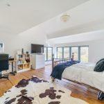 Property for sale by Auction A bright bedroom features a large window with scenic views, a bed with metal frame and white linens, a cowhide rug on wooden floors, and a workspace with a desk, computer, and office chair. Opposite the bed is a white dresser with a TV on top and storage shelves. The decor hints at rustic charm from Hagg Hill Farm. Presented by Palace Auctions