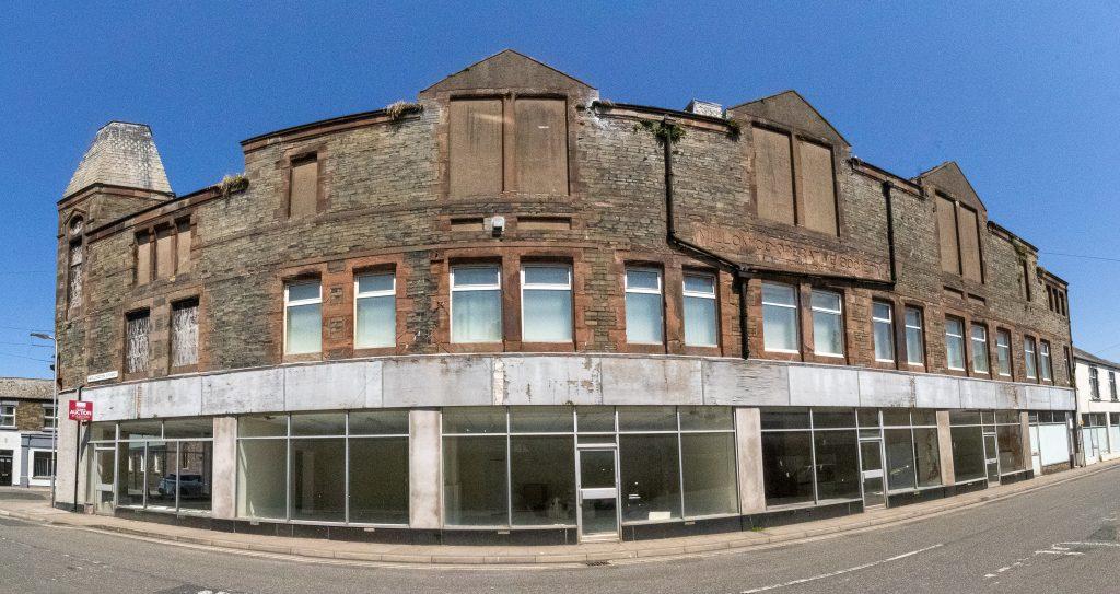 Property for sale by Auction A wide-angle view of a large abandoned building with boarded-up windows and dilapidated brickwork. The structure is on a street corner and has a row of empty storefronts with peeling paint and broken glass. The clear blue sky contrasts with the building's worn appearance. Presented by Palace Auctions
