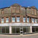 Property for sale by Auction A wide-angle view of a large abandoned building with boarded-up windows and dilapidated brickwork. The structure is on a street corner and has a row of empty storefronts with peeling paint and broken glass. The clear blue sky contrasts with the building's worn appearance. Presented by Palace Auctions