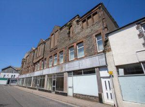 Property for sale by Auction A row of old, abandoned buildings with boarded-up windows and faded signs under a clear blue sky. The structures are primarily made of stone and brick, showing signs of decay. There are no people or vehicles in sight. The scene evokes a sense of desolation and neglect. Presented by Palace Auctions