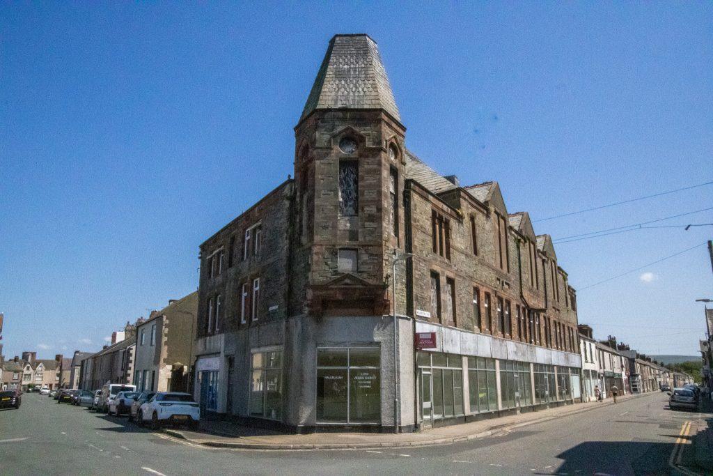 Property for sale by Auction A street corner featuring a large, historic, multi-story stone building with an angled, turreted roof. The building has large windows on the ground level, some covered with white shades. Parked cars line the street on a bright, sunny day under a clear blue sky. Presented by Palace Auctions