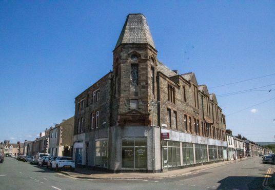 Property for sale by Auction A street corner featuring a large, historic, multi-story stone building with an angled, turreted roof. The building has large windows on the ground level, some covered with white shades. Parked cars line the street on a bright, sunny day under a clear blue sky. Presented by Palace Auctions