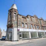 Property for sale by Auction An old, abandoned building made of stone with boarded-up windows stands under a clear, blue sky. The structure has a prominent tower on one corner and a row of large glass windows on the bottom floor. Next to it is another building with a peaked roof. Presented by Palace Auctions