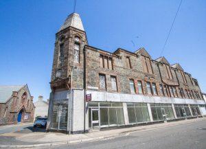 Property for sale by Auction An old, abandoned building made of stone with boarded-up windows stands under a clear, blue sky. The structure has a prominent tower on one corner and a row of large glass windows on the bottom floor. Next to it is another building with a peaked roof. Presented by Palace Auctions