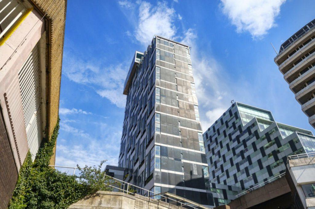 Property for sale by Auction View from the ground of three modern high-rise buildings with varying architectural styles against a bright blue sky with scattered clouds. One building has ivy climbing its wall, creating a lush facade. The scene, which could be home to a Liverpool penthouse investment apartment, includes a concrete structure in the foreground. Presented by Palace Auctions