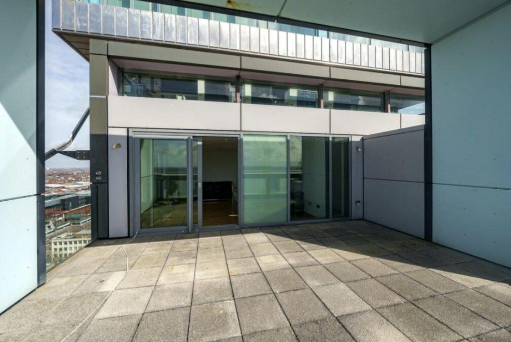 Property for sale by Auction A spacious balcony with a cement floor offers views of the city. Glass doors and windows lead to the interior of a modern Liverpool penthouse investment apartment. Metal and glass structures surround the area, providing a sleek and contemporary feel. The sky is clear in the background. Presented by Palace Auctions