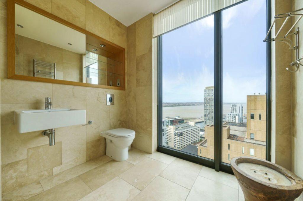 Property for sale by Auction A modern bathroom in a Liverpool penthouse investment apartment, featuring beige marble walls and floors, a floating sink, toilet, and large window showcasing cityscape views with buildings and a river. The room includes built-in shelves with a mirror and a rustic bowl-style basin. Presented by Palace Auctions