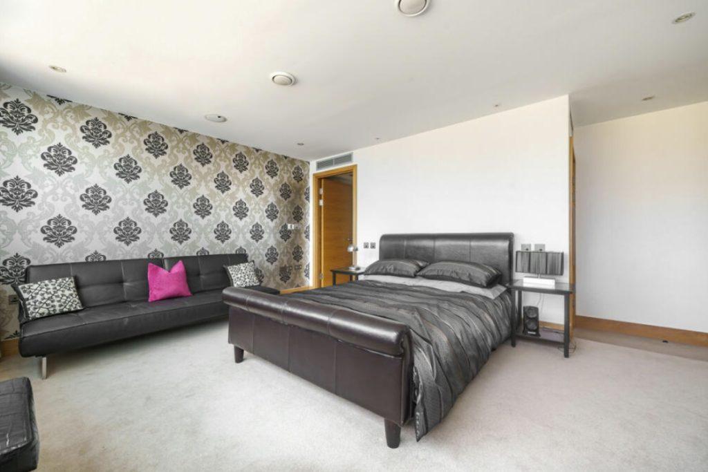 Property for sale by Auction A modern Liverpool penthouse investment apartment features a bedroom with a leather bed frame, grey bedding, and black nightstands. A black sofa with decorative pillows and a pink accent pillow sits against a wall adorned with black and silver damask wallpaper. The room boasts light carpeting and recessed lighting. Presented by Palace Auctions