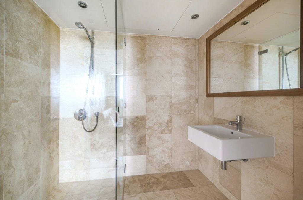 Property for sale by Auction A modern bathroom in a Liverpool penthouse investment apartment with beige tile walls and floors. It features a glass-enclosed shower with a wall-mounted shower head on the left. On the right, there is a rectangular white sink with a silver faucet and a large mirror above it. Presented by Palace Auctions
