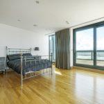 Property for sale by Auction A spacious, modern bedroom in a Liverpool penthouse investment apartment, featuring wooden flooring and a metal-frame bed with dark bedding. An ornate bedside table with a lamp and a large floor-to-ceiling window opens to a balcony with a scenic view. A gray chair is positioned near the window. Presented by Palace Auctions