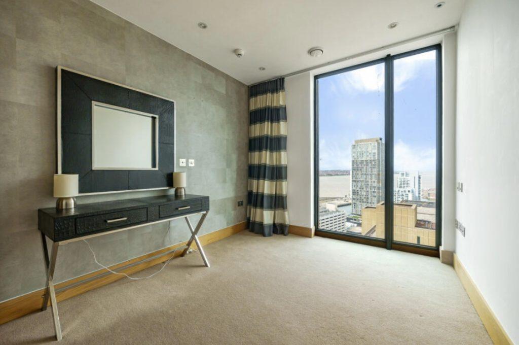 Property for sale by Auction A minimalist room in a Liverpool penthouse investment apartment boasts a large window offering city and river views. The room features a modern table with two lamps, a framed mirror on the wall, striped curtains, light wooden accents, and neutral-colored walls. Presented by Palace Auctions