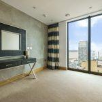 Property for sale by Auction A minimalist room in a Liverpool penthouse investment apartment boasts a large window offering city and river views. The room features a modern table with two lamps, a framed mirror on the wall, striped curtains, light wooden accents, and neutral-colored walls. Presented by Palace Auctions