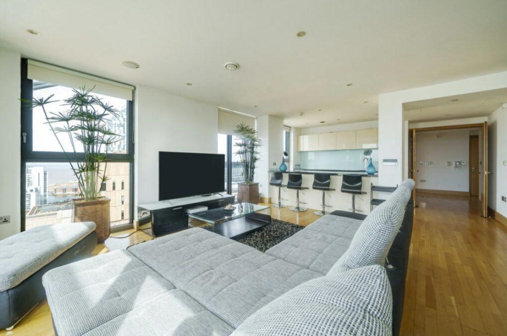 Property for sale by Auction A Liverpool penthouse investment apartment features a modern living room with light wood flooring, a large gray sectional sofa, and a flat-screen TV on a sleek black stand. Tall plants are beside the windows, and a kitchen with white cabinets and a breakfast bar with black high chairs is visible in the background. Presented by Palace Auctions