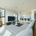 Property for sale by Auction A Liverpool penthouse investment apartment features a modern living room with light wood flooring, a large gray sectional sofa, and a flat-screen TV on a sleek black stand. Tall plants are beside the windows, and a kitchen with white cabinets and a breakfast bar with black high chairs is visible in the background. Presented by Palace Auctions