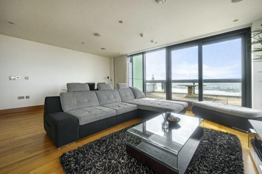 Property for sale by Auction A modern living room in a Liverpool penthouse investment apartment features a large gray sectional sofa and matching ottoman on a black shag rug. A glass-top coffee table sits in front of the sofa, while floor-to-ceiling windows and doors offer an expansive view of the balcony and cityscape beyond. Presented by Palace Auctions